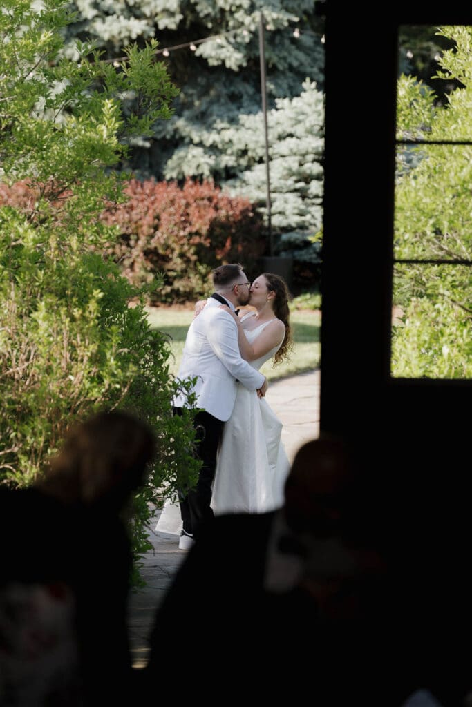 A travel wedding photographer captures a couple as they sneak in a kiss outside their wedding ceremony at Wildflower on Watts in Honesdale PA surrounded by flowers.