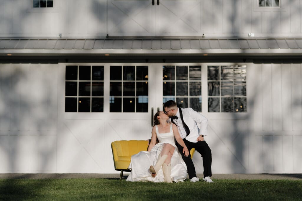 A newly married Bride and Groom sit and kiss each other on a yellow couch outside the big white barn at Wildflower on Watts in Honesdale PS.