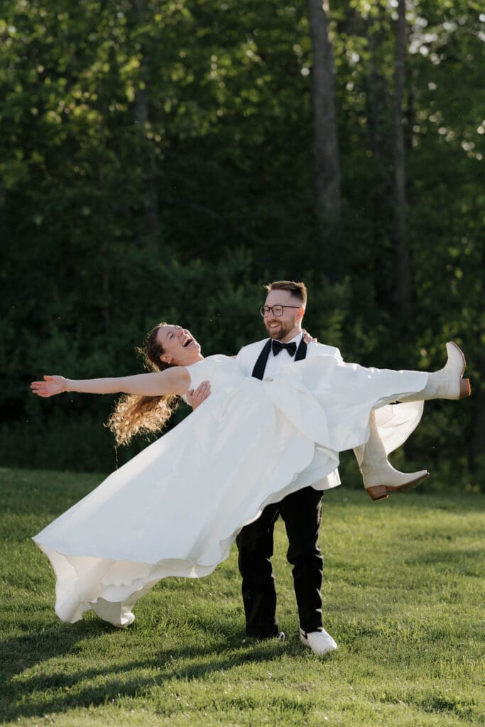 A Bride and Groom celebrate following their wedding ceremony. She is in his arms laughing with her white cowboy boots kicked up, as they both smile at each other lovingly. Captured by travel wedding photographer Hayley Zumkeller photography and videography team from Victoria BC.