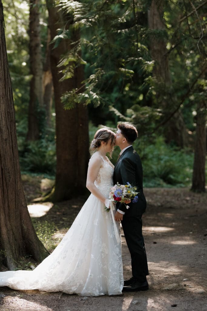 A newly married bride and groom have photos captured in a private wooded area with sun rays streaming through the trees. He is kissing her forehead as they embrace.