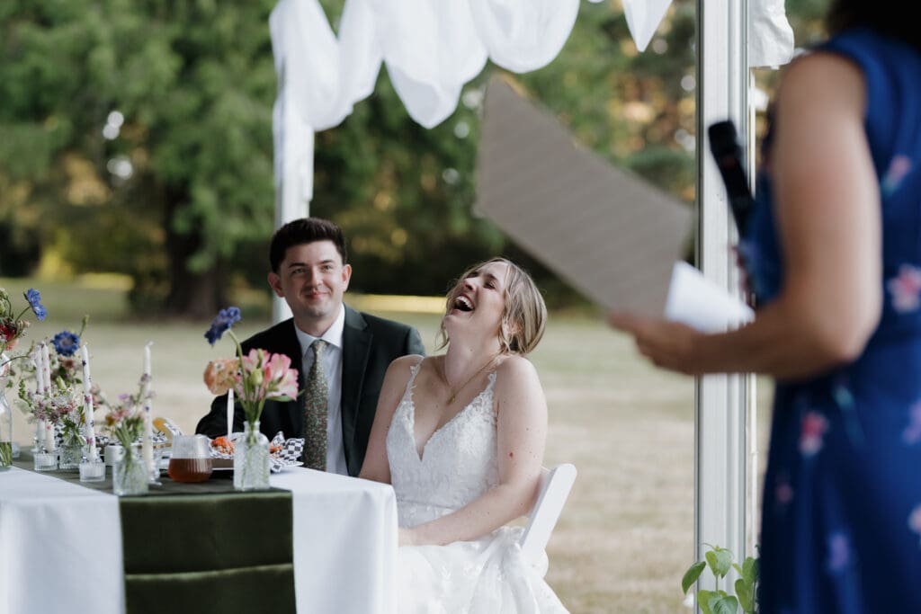A bride and groom laugh during speeches at their outdoor wedding ceremony in Nanaimo BC. Captured by Hayley Zumkeller's Vancouver Island Wedding Videography team.