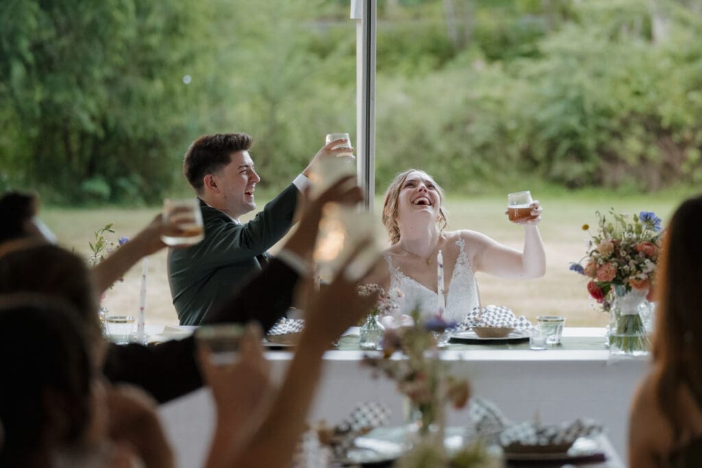 Everyone raises a glass as we see the bride and groom laughing during outdoor wedding speeches at their dinner. Captured by Hayley Zumkeller Photography and Videography team in Nanaimo BC.