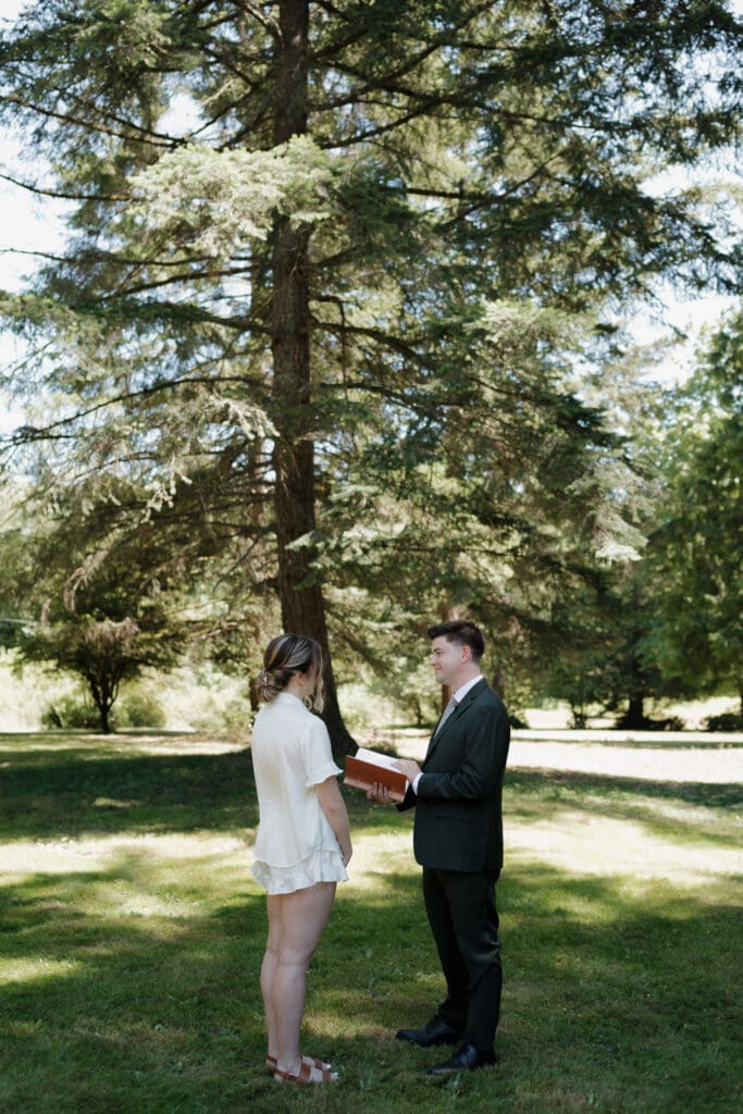 A bride and groom exchange private vows and letters during their intimate first look among the trees outlining the family property location of the ceremony in Nanaimo BC. The bride wears white bridal pyjamas while the groom is fully dressed in his wedding attire.
