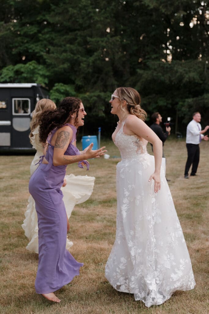 A bride and her friend (dressed in a lilac dress) laugh and dance during the outdoor wedding reception. A food truck can be seen in the background.