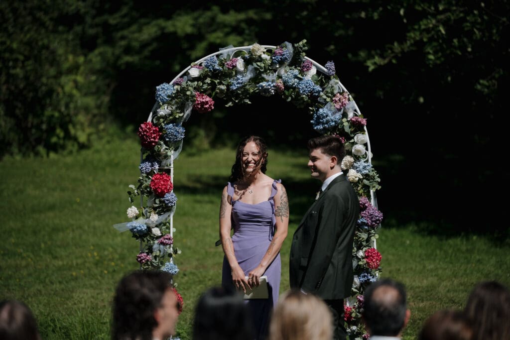 A friend of the bride and groom dressed in a lilac dress stands under a floral arch at the front of the outdoor ceremony ready to be a celebrant and lead this special event. The groom is to her left as they await the arrival of the bride.