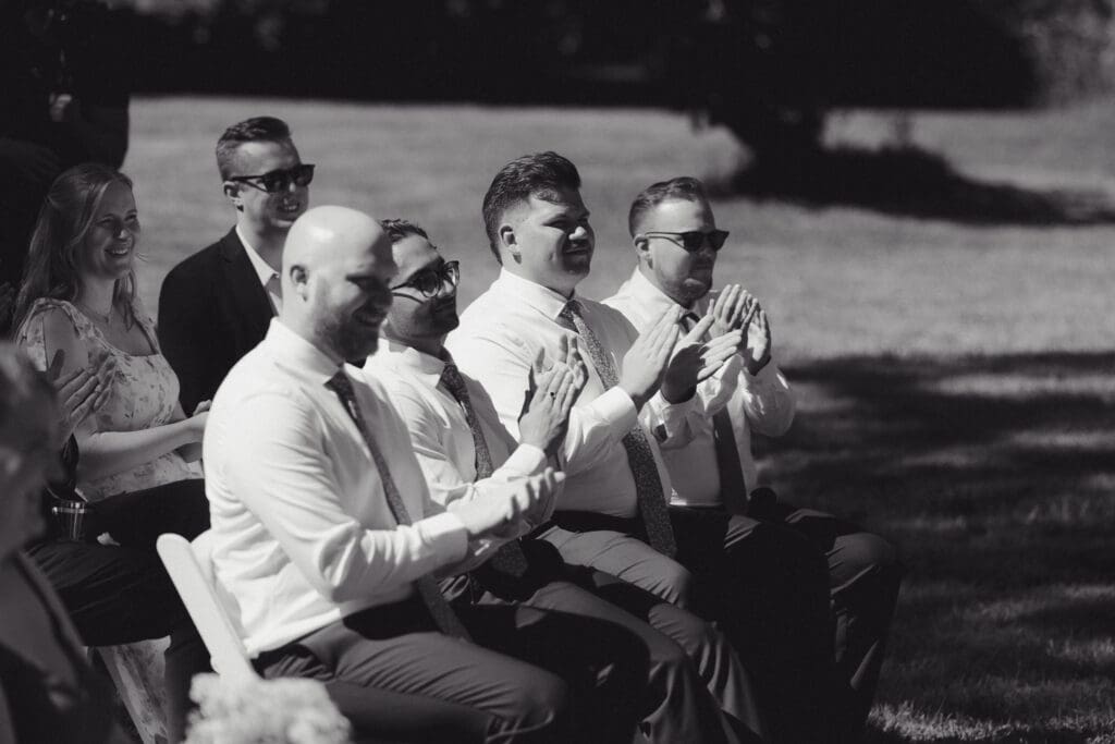 The groomsmen of a wedding are sitting and clapping following the ceremony in this black and white image. Captured by Hayley Zumkeller's Vancouver Island Wedding Videography team.