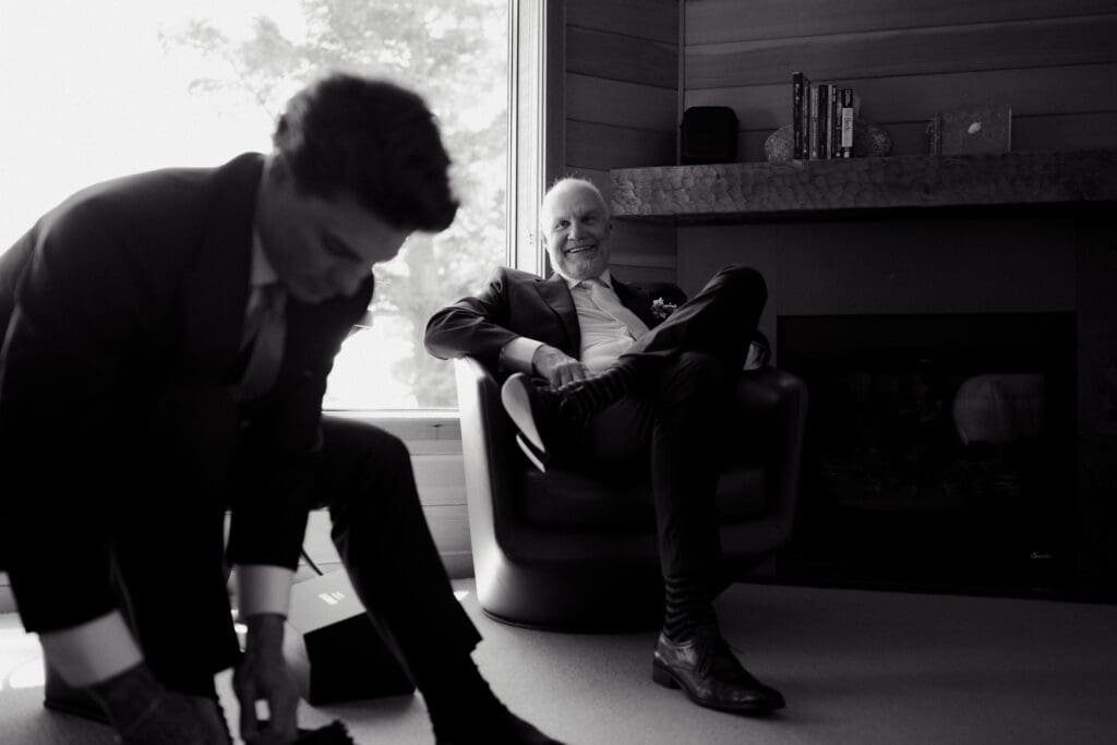 A black and white image of a groom getting ready by tying his shoes before his wedding with his father in the background watching on proudly.