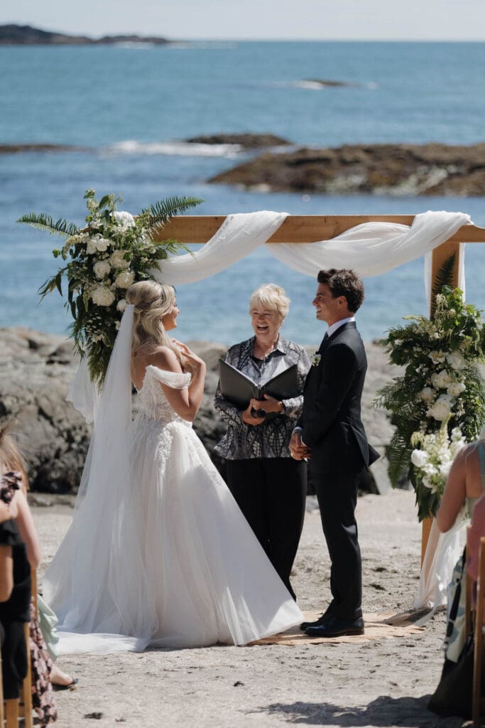 A shot of the bride and groom at the front with their guests behind during a private beach wedding ceremony at the Wickaninnish Inn on Vancouver Island. Ocean and rocks behind them.