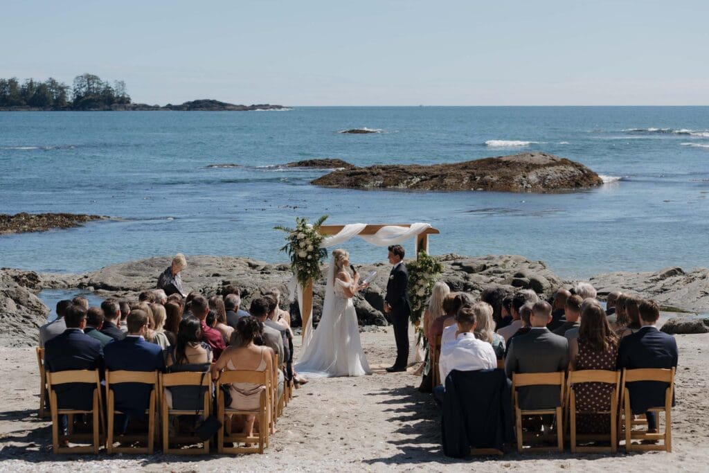 A shot of the bride and groom at the front with their guests behind during a private beach wedding ceremony at the Wickaninnish Inn on Vancouver Island. Ocean and rocks behind them.