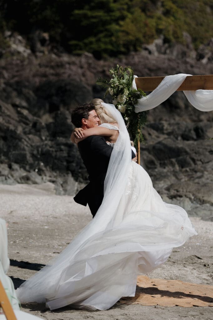 A newly married couple embracing after their private beach ceremony on Chesterman Beach. The groom is lifting the bride as her cathedral-length veil wraps around them.