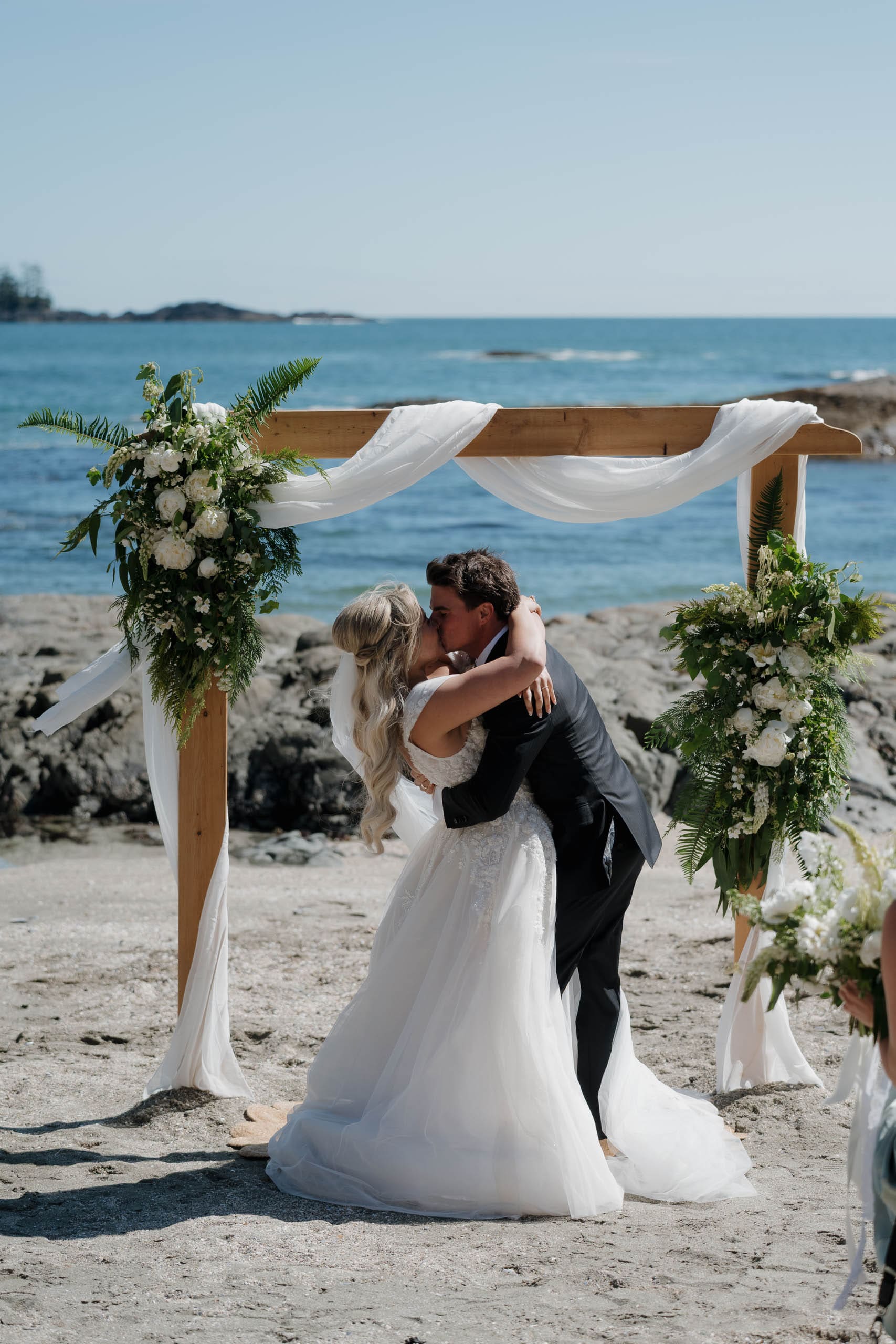 Tofino wedding photographer captures a bride and groom sharing their first kiss as husband and wife at their beach wedding, with the Pacific Ocean in the background at Chesterman Beach.
