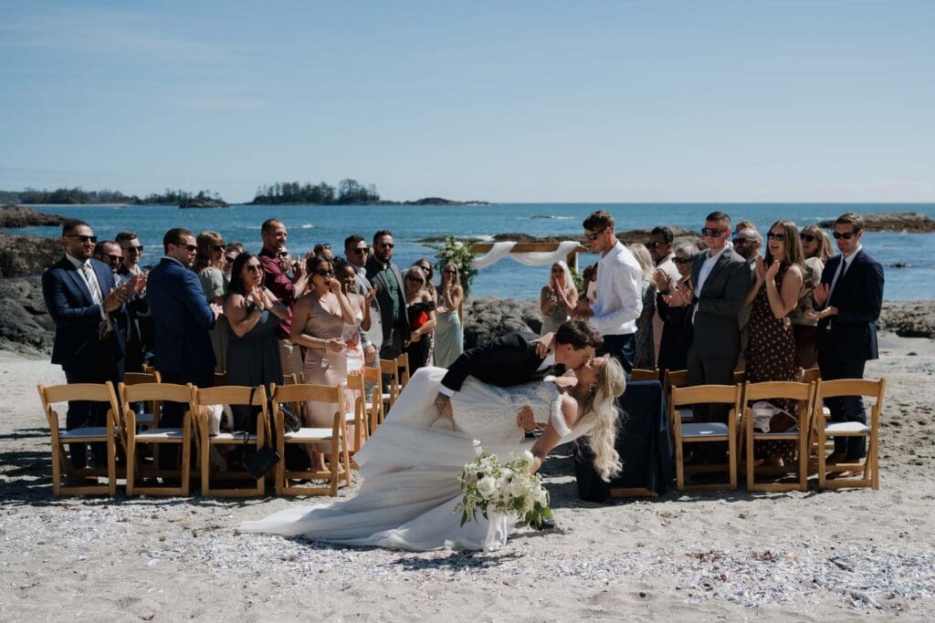 A groom dip kisses his bride as they exit the ceremony with all the guests cheering in the background of this beach side wedding in Tofino BC.