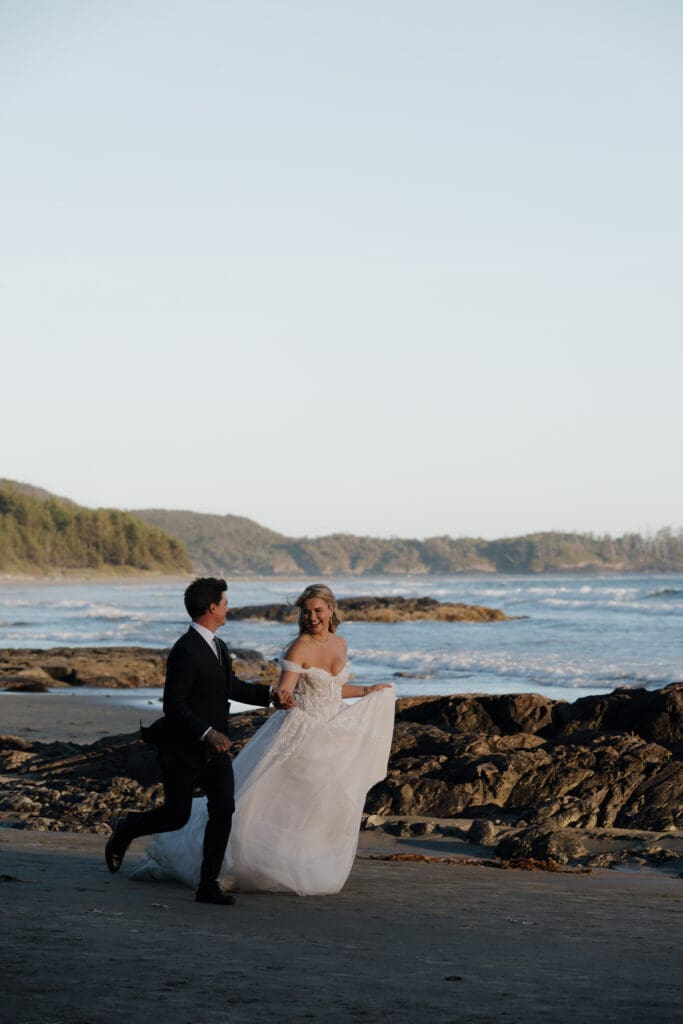 June wedding photos in Tofino BC by a Tofino wedding photographer at the Wickaninnish Inn shows a newly married bride and groom running along Chesterman Beach with the ocean and rock formations in the background.