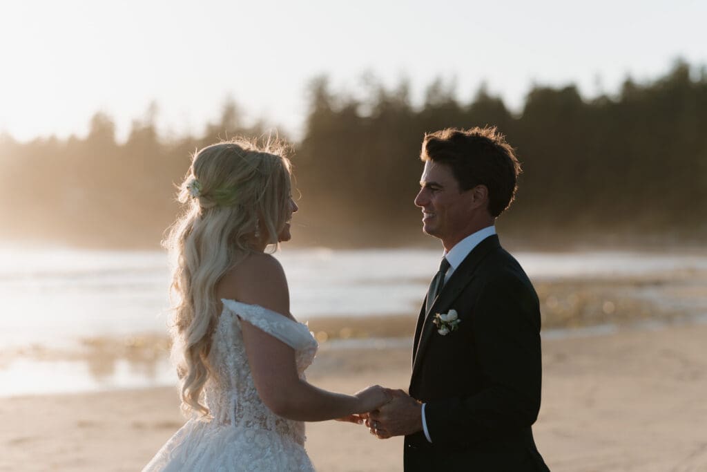 Newlyweds embrace hands and stare into each others eyes during golden hour sunset with the Pacific Ocean behind them in Tofino BC