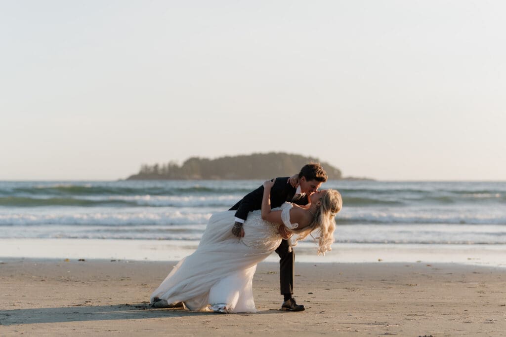 A bride and groom on the beach with the setting golden hour sun are engaged in a deep dip kiss with the ocean waves behind them.