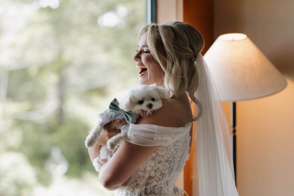 A bride holds her pet dog as she laughs while looking out the window, just following getting ready at Wickaninnish Inn.