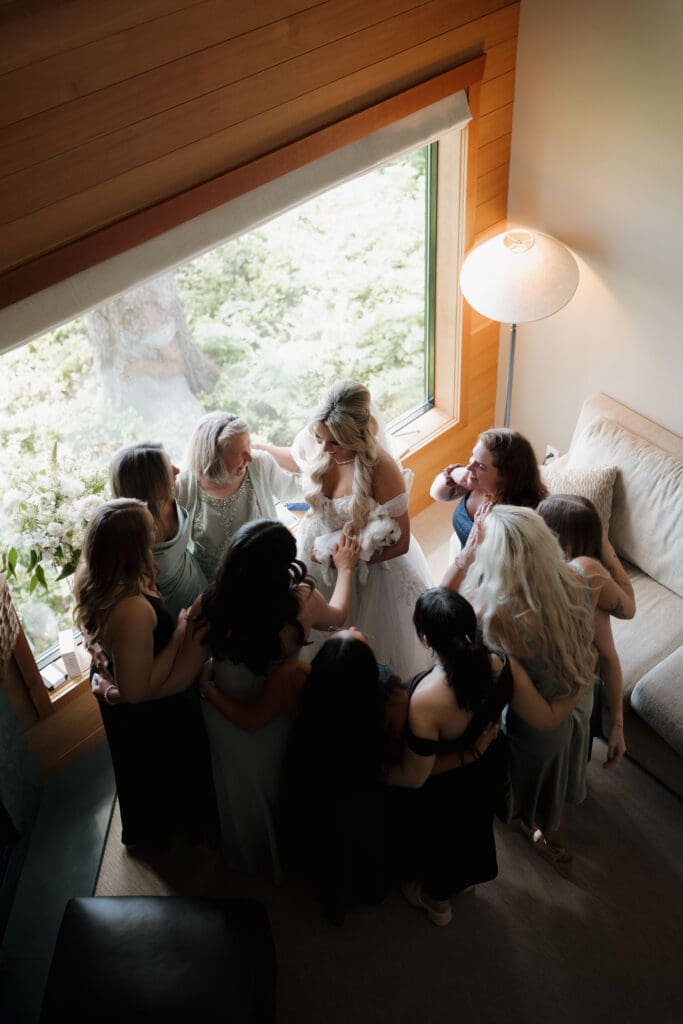 An overhead shot of a Bride and bridesmaids getting ready in an oceanfront suite at the Wickaninnish Inn in Tofino BC following their first look at her fully dressed in her gown, exclaiming happily and smiling.