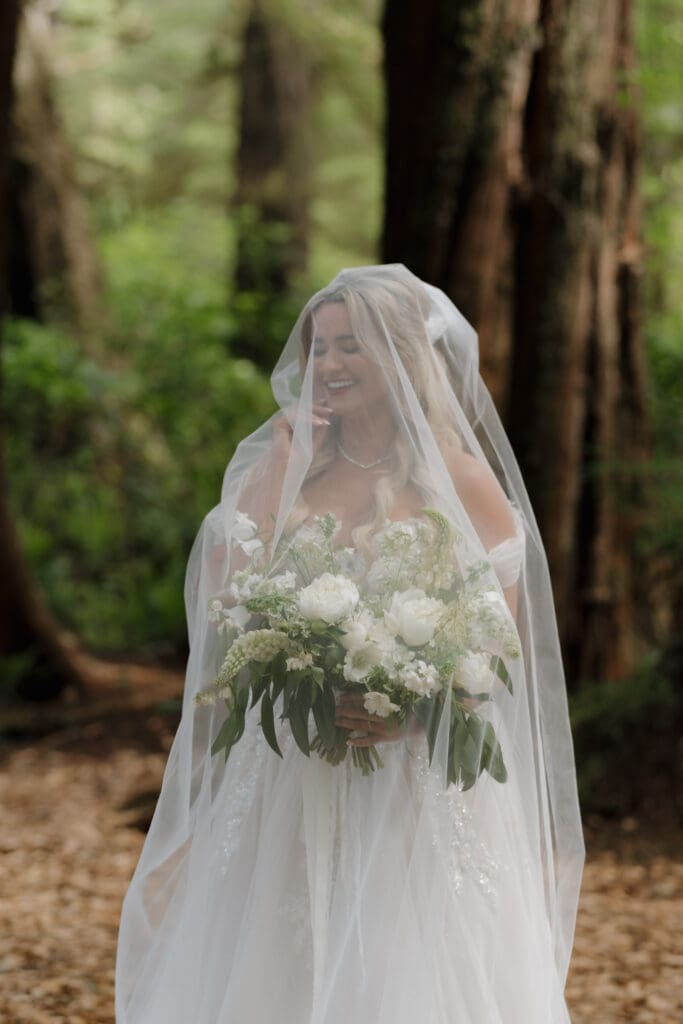 A bride in the rainforest of Tofino BC with her veil gently shielding her face as the light streams through the trees.