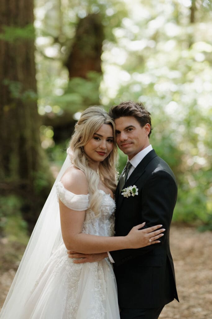 A bride and groom embrace and pose for the camera following their first look as the light filters through the rainforest tree of Tofino BC.