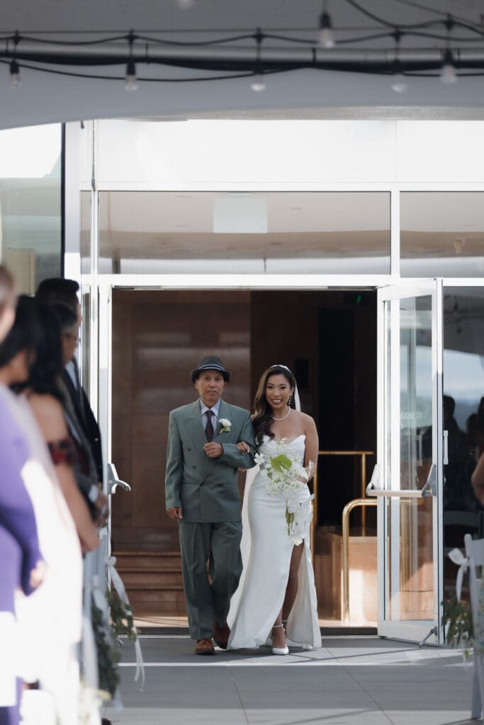 A bride and her dad walk through glass doors to head down the aisle during the wedding ceremony at Inn on Laurel Point in Victoria BC. We also see guests on either side turned to look at them.