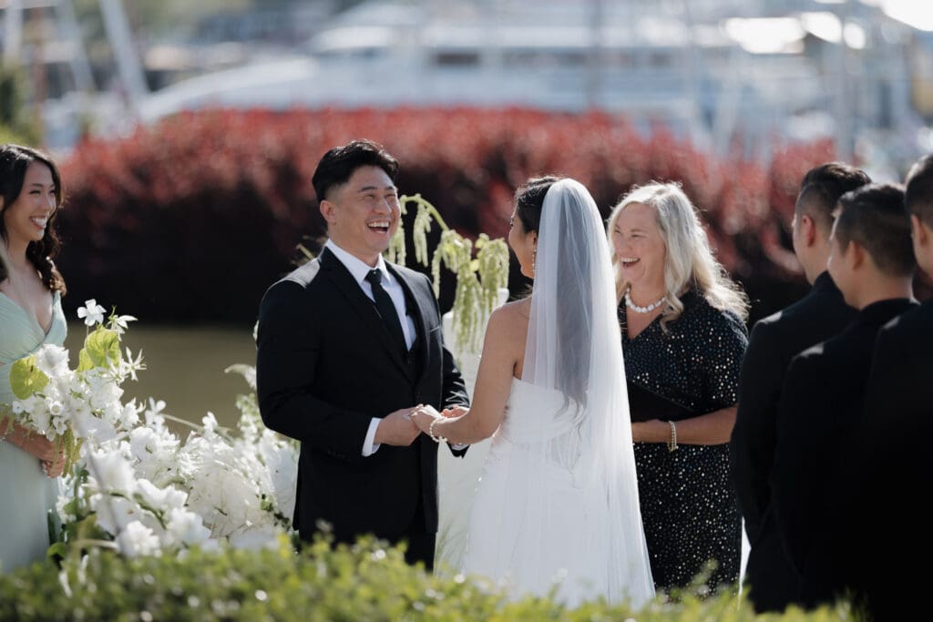 A laughing groom is the primary focus of this Inn at Laurel Point Wedding ceremony photo in Victoria BC. He is holding his bride's hands and we see her beautiful veil over her dark hair. The officiant is also laughing.