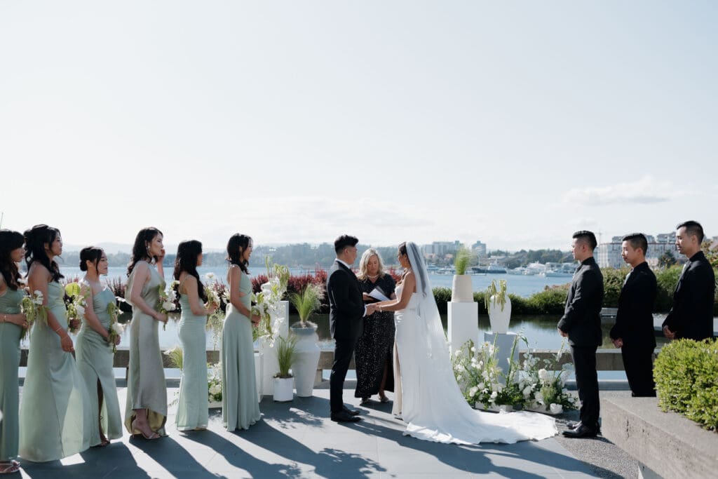 An image of a wedding ceremony held at Inn at Laurel Point in Victoria BC with the bride and groom exchanging vows. Bridesmaids and groomsmen on either side. And the harbour front behind them.